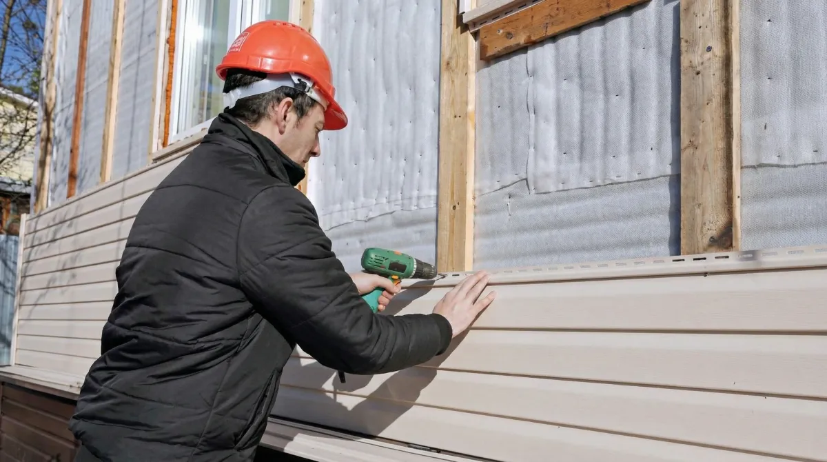 Siding replacement professional using a power drill to install new tan vinyl siding on a home exterior in Newark, NJ